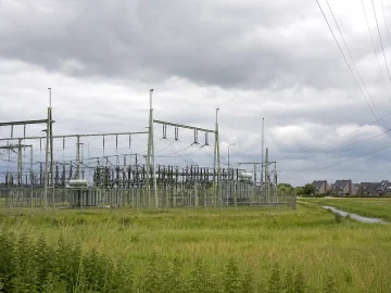Electricity substation equipment and high-voltage power lines beside grassy fields under a cloudy sky.