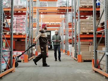 A warehouse worker pulls a loaded cart down an aisle between tall racks