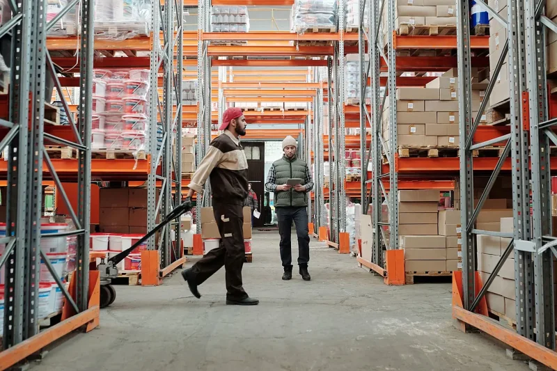 A warehouse worker pulls a loaded cart down an aisle between tall racks