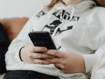 A teenager in a white hoodie sits on a sofa holding a smartphone, face out of frame